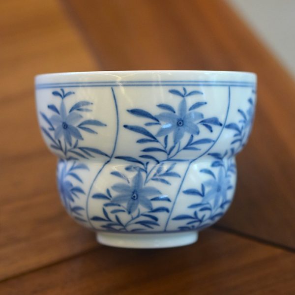 Close-up of a blue and white porcelain gourd coffee cup with hand-painted floral pattern: two six-petal flowers among leaves, natural light on brown wooden background.