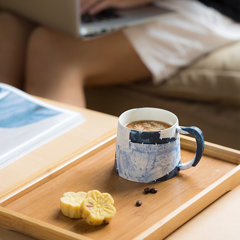 A handmade ceramic coffee mug on a wooden table, with a person working in the background on a cozy sofa.