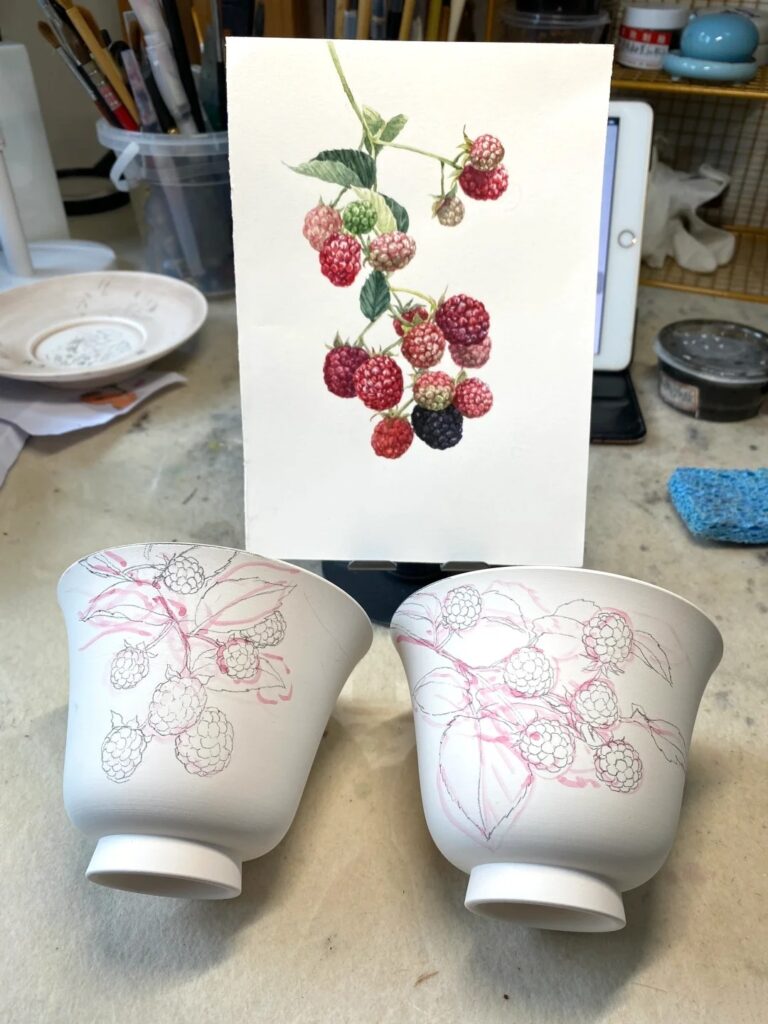 Artist hand-painting floral design onto a ceramic cup using original sketch as reference